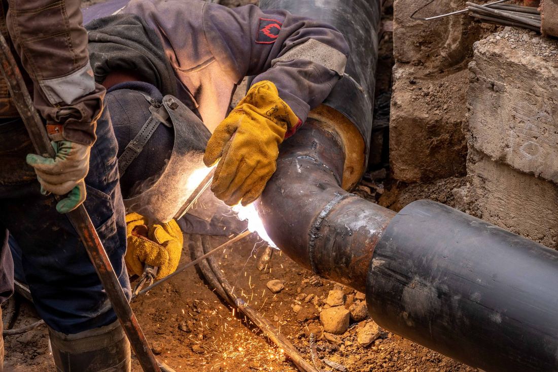 A tradesman welding a pipe in a trench with both hands occupied, unable to reach a ringing phone.